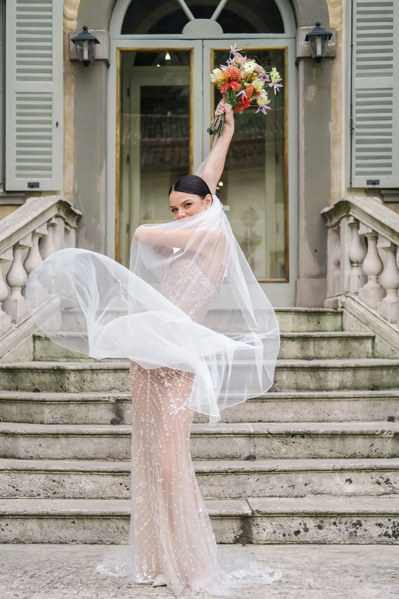 Mariée élégante en robe de mariage avec voile en mouvement, tenant un bouquet de fleurs colorées sur les marches de la Villa Canton à Milan.