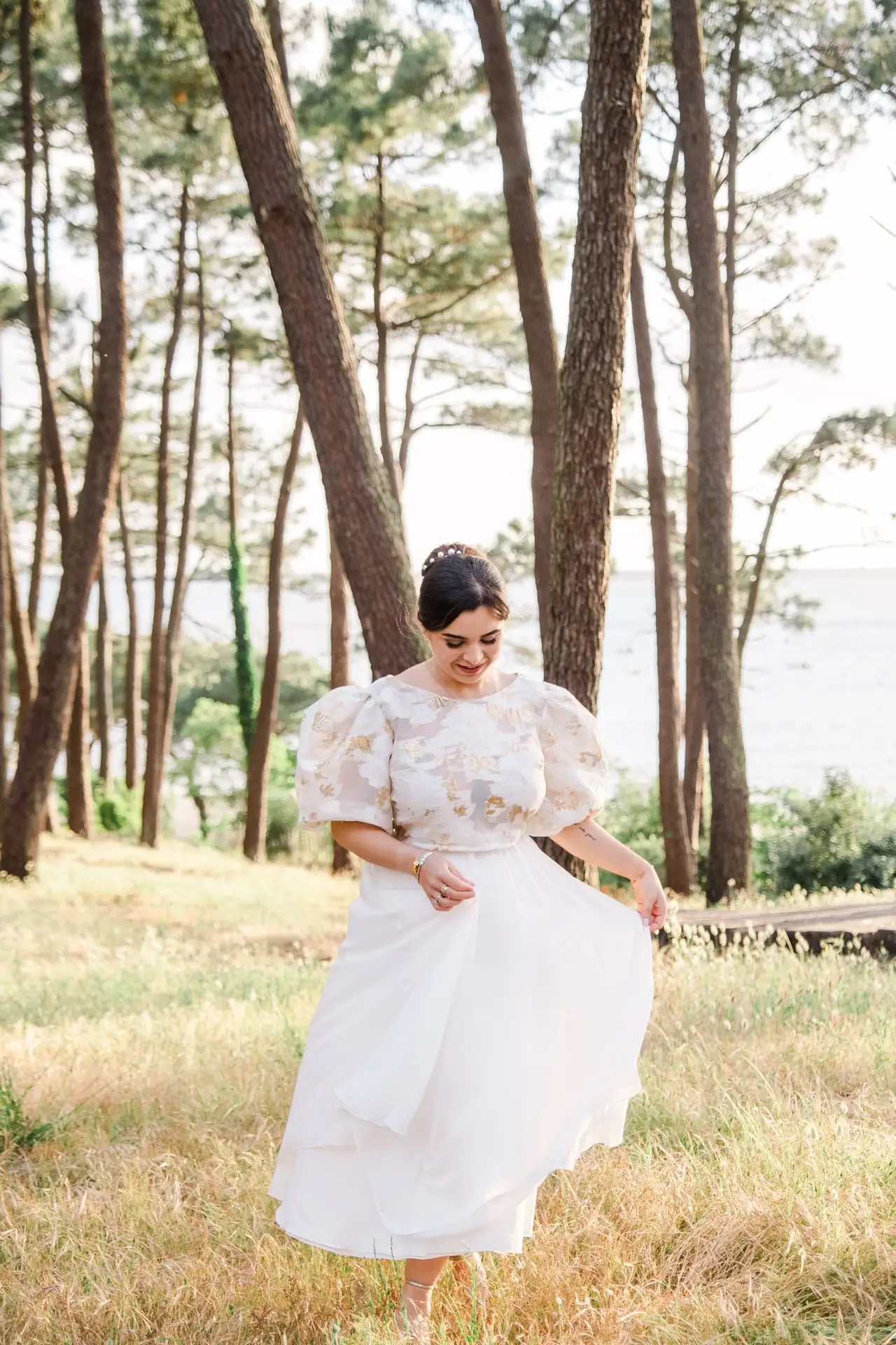 Femme en robe de mariage fluide marchant dans la nature sur le Bassin d’Arcachon, entourée de pins, lumière douce et ambiance poétique.