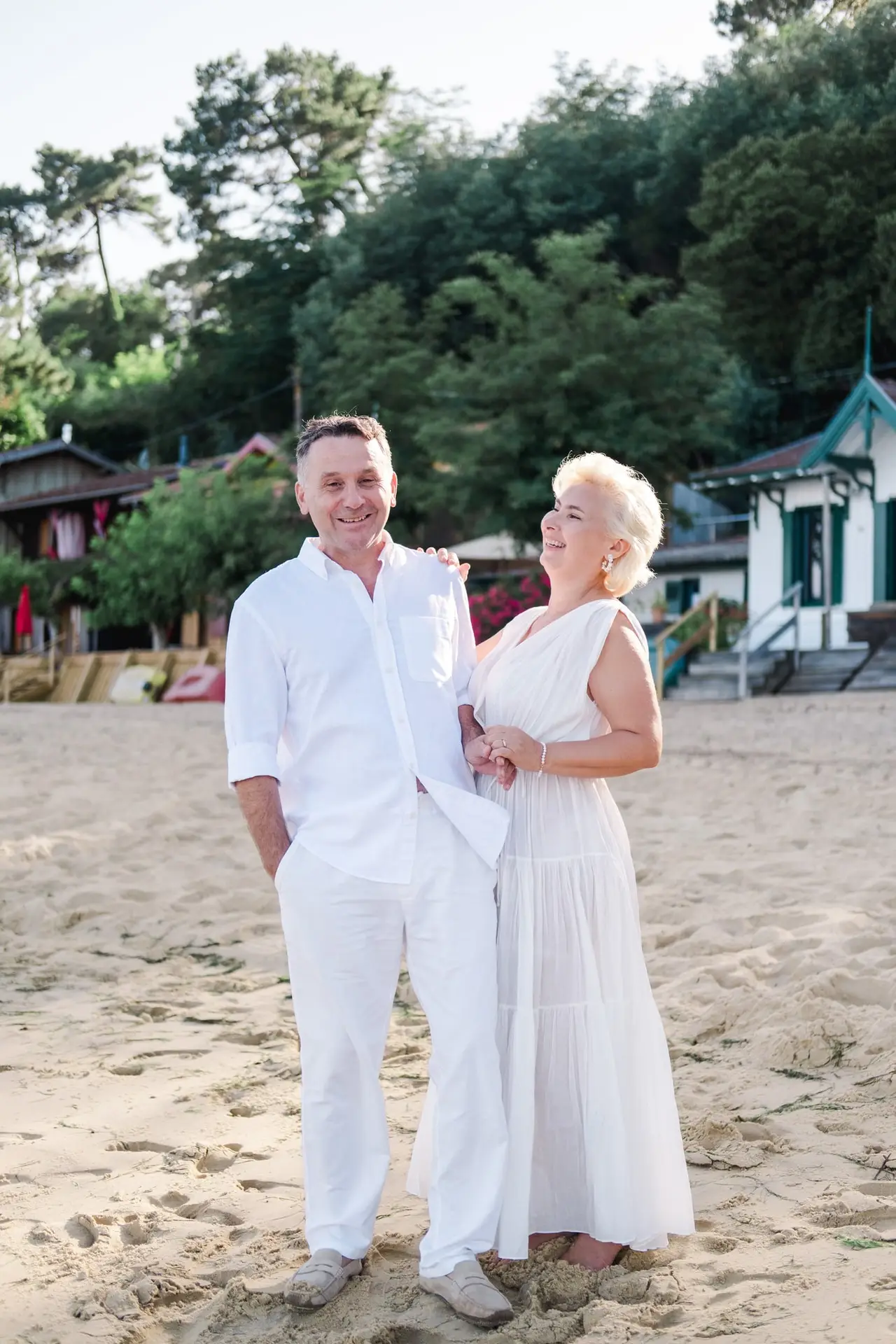 Couple souriant vêtu de blanc sur la plage du Cap Ferret, dans le village de L’Herbe, partageant un moment complice dans une ambiance lumineuse et naturelle.