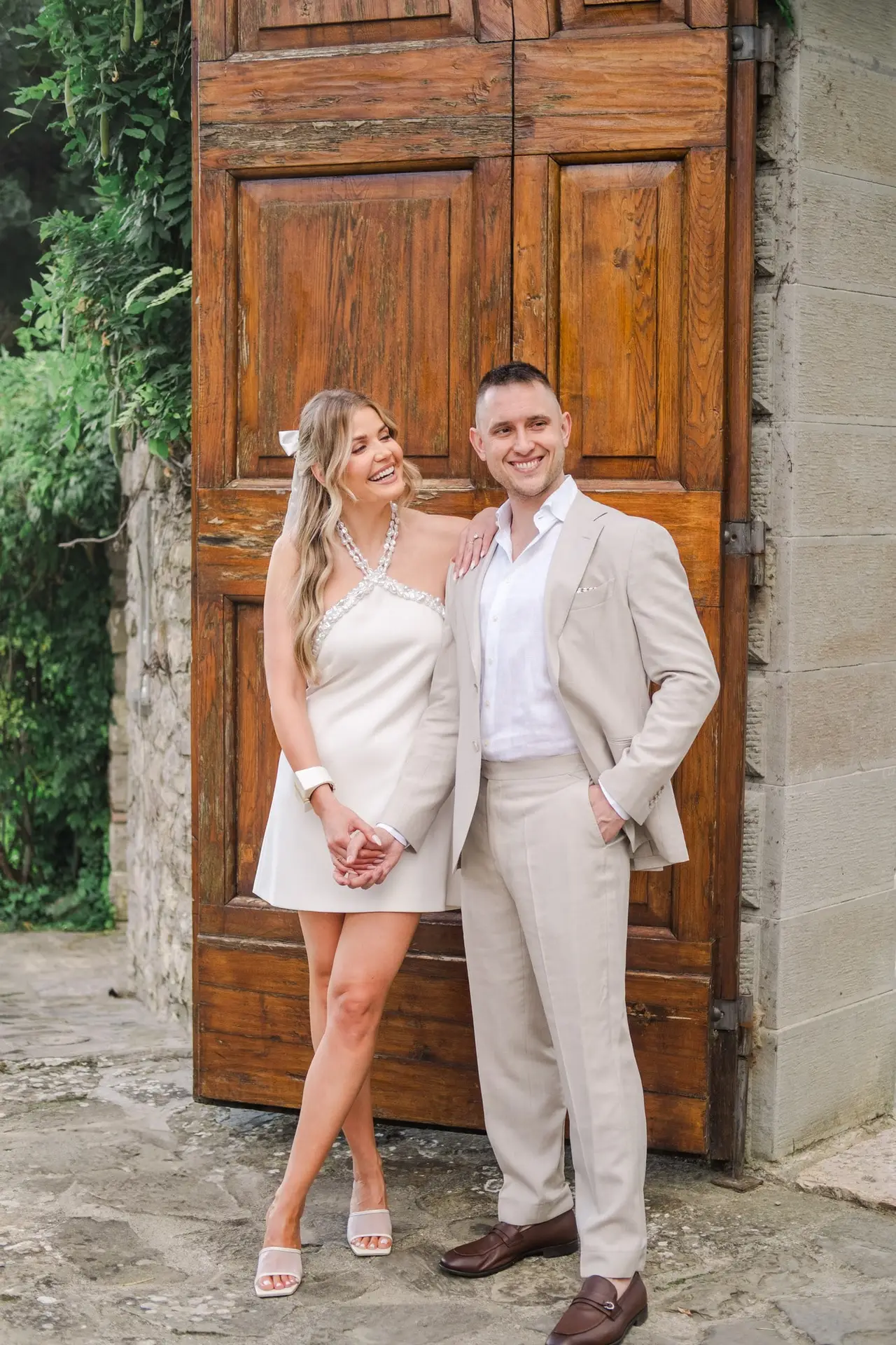 Couple élégant en tenue claire se tenant la main devant une porte en bois au Borgo Corsignano en Toscane, photographié par Julien Boyer.