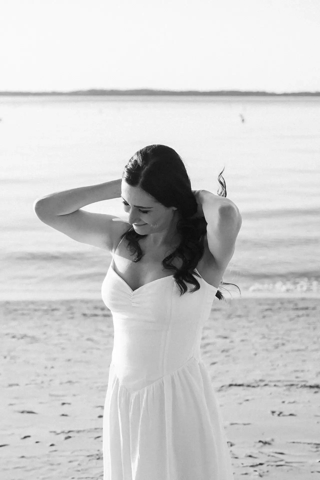 Portrait en noir et blanc d’une femme en robe claire sur la plage, regard doux et posture naturelle face à la mer.